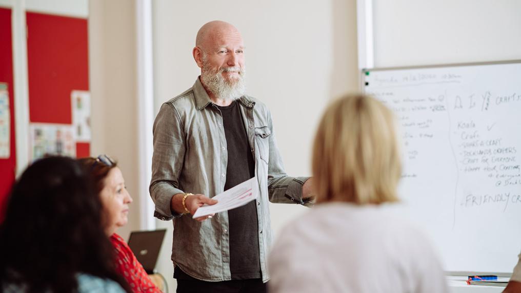 Older white man with beard stands talking and smiling in a workshop setting