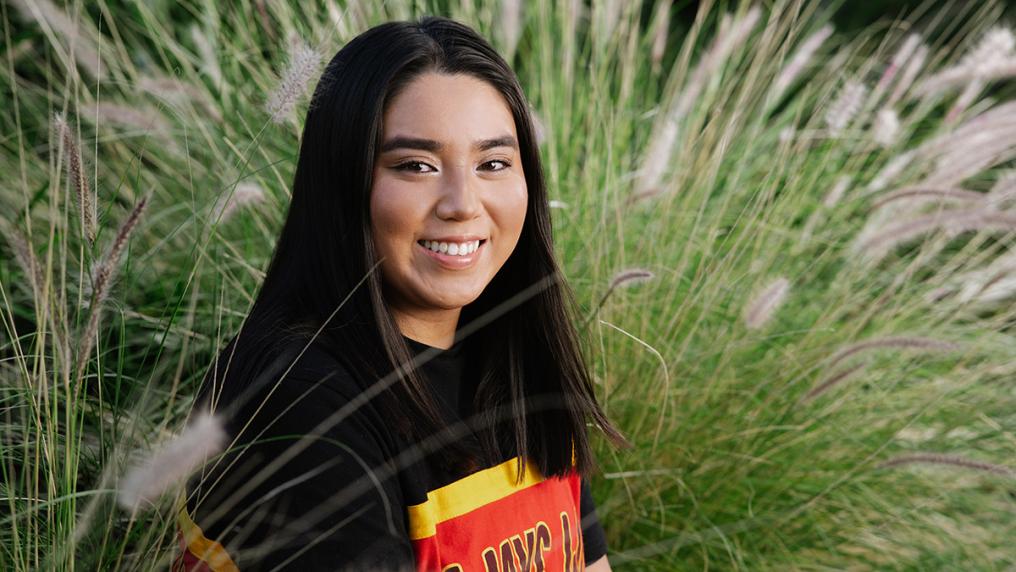 Young woman in a T-Shirt with the Aboriginal flag in front of a large green plant, smiling