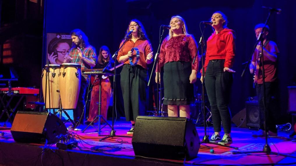A group of young musicians perform on stage, with 3 singers, a drummer and 2 guitarists
