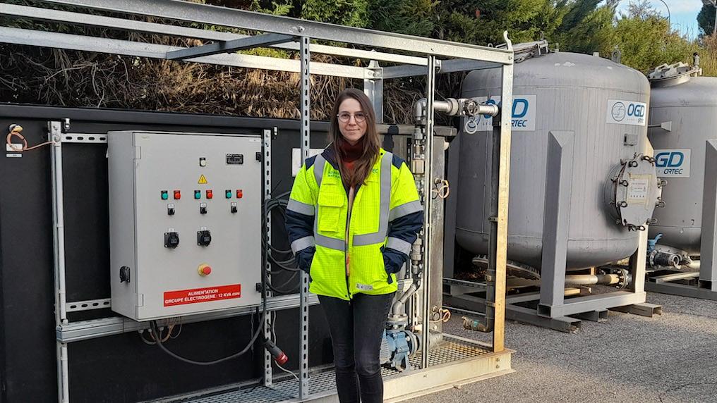 A young woman in a high-visibility jacket stands near water tanks and electronic equipment