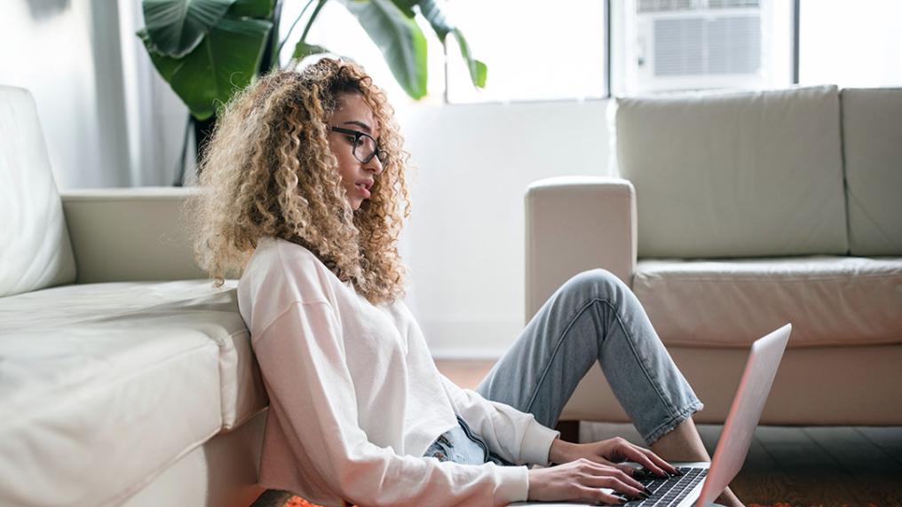 A student at home sits on the floor holding a laptop.