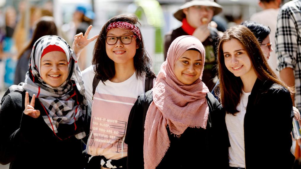 Four students standing together outside, looking and the camera and smiling. Event stalls and people walking around in the background, out of focus.