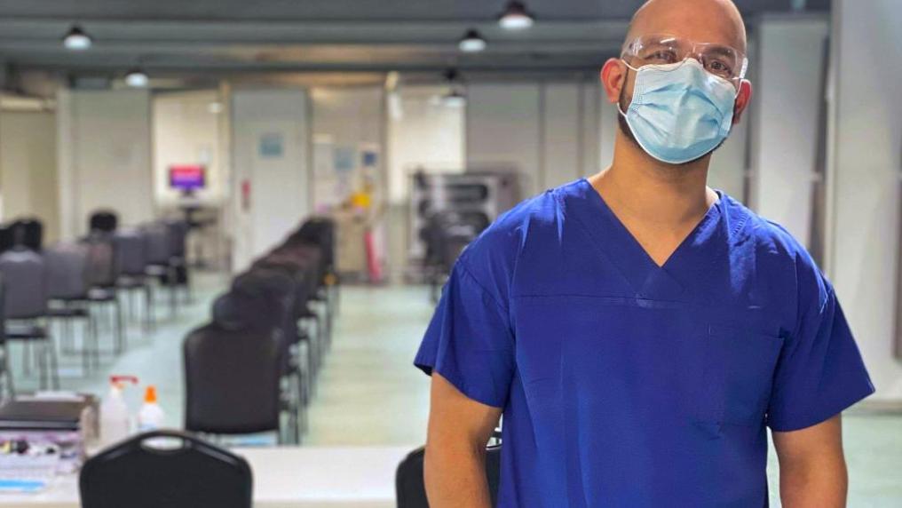 A male in nurse's uniform wears a mask and protective glasses, in a large white room with rows of chairs