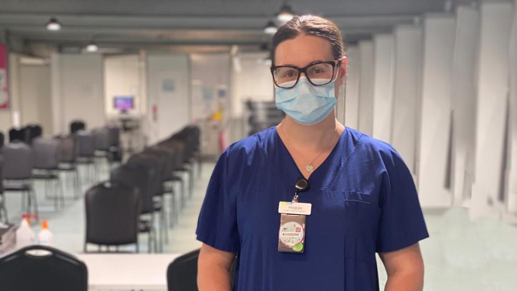 A young white woman in a nurse's uniform, surgical mask and glasses stands in a large white room with rows of chairs