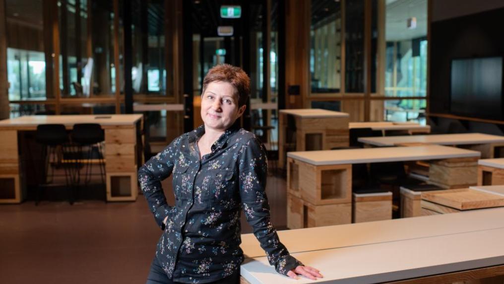 Person looking at the camera while standing in an empty classroom, leaning on one of the desks