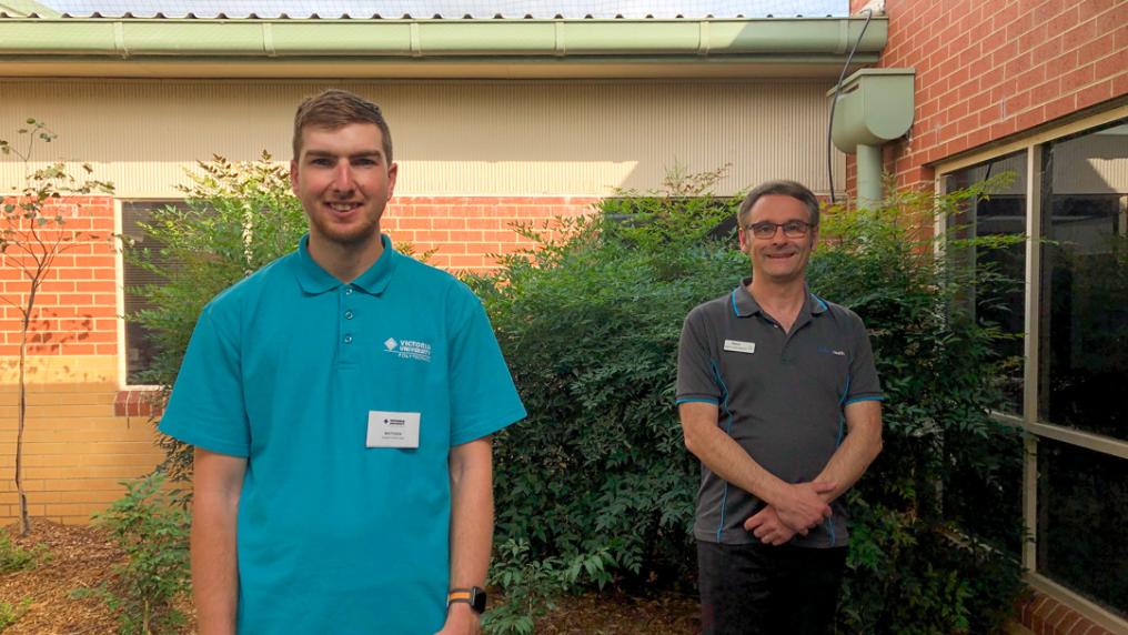 Two people standing outside and smiling at camera. One wearing a blue Victoria University shirt, the other wearing a dark Northern Health shirt.