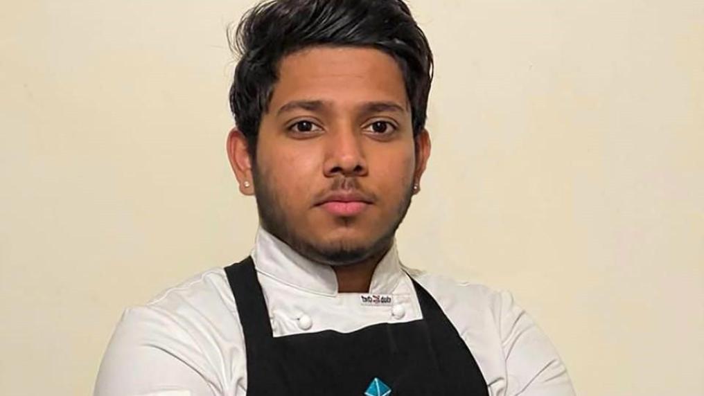 Pasan wearing a chef's uniform and apron, standing in front of a plain background.