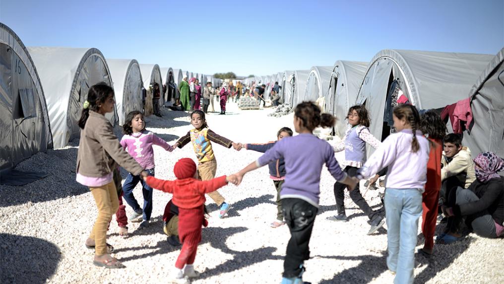 A group of children hold hands in a circle in what looks like a game, in an arid tent community