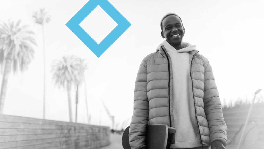 A smiling student holding a skateboard stands next to the VU branded blue diamond. 