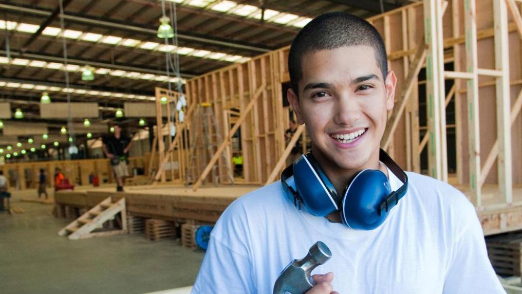 A carpentry student with ear protection around their neck. The student is holding a hammer. In the background is a house frame being built.