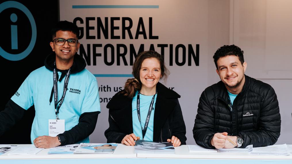 Three young adults in blue t-shirts with the Victoria University logo smile in front of a sign that reads 'General information' 