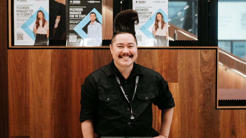 A man with his hair in a swept up wave smiles in front of Victoria University signage