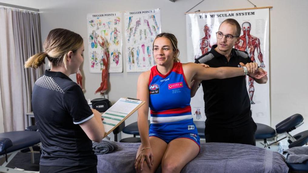 A person in a Western Bulldogs uniform having their arm streteched by a VU student. Another person is holding a clipboard and reading it.