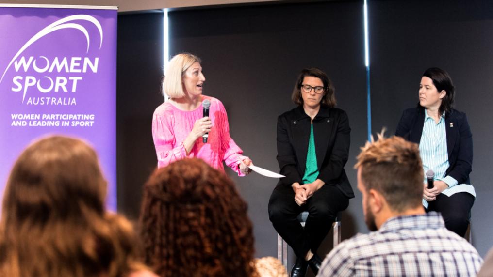 Three people on a stage, one holding a microphone and talking. To the left is a banner that says 'Women Sport Australia'