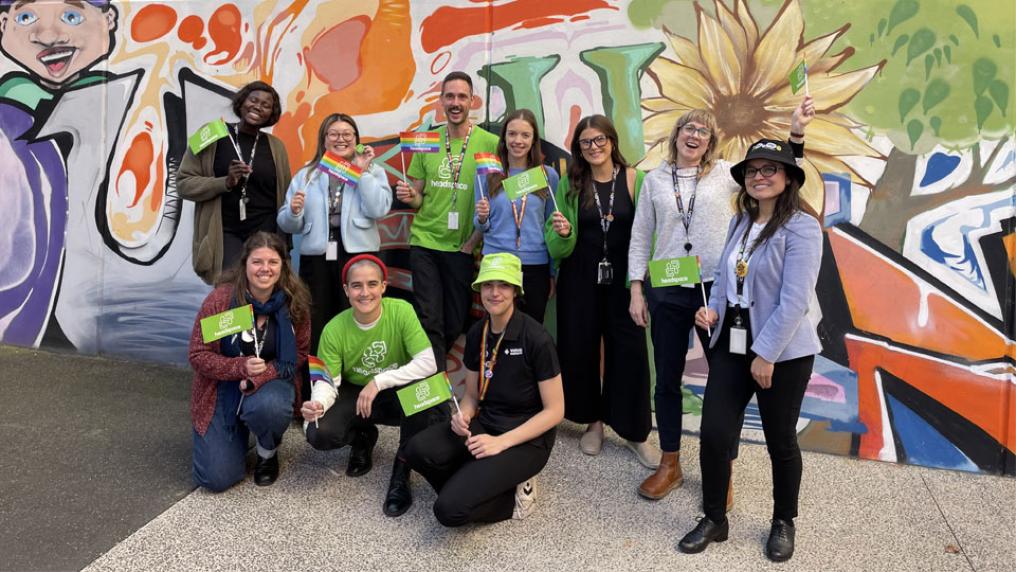 Briahny with the team at headspace Sunshine. The team are standing outside a building with colourful street art, smiling and waving rainbow and headspace flags. 