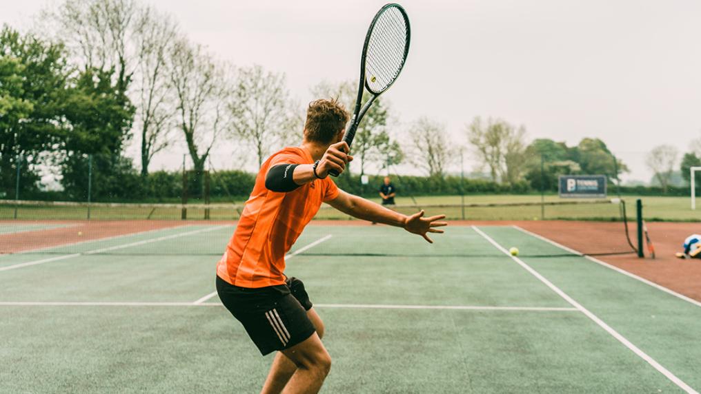 A man in orange sport uniform plays tennis on an asphalt court