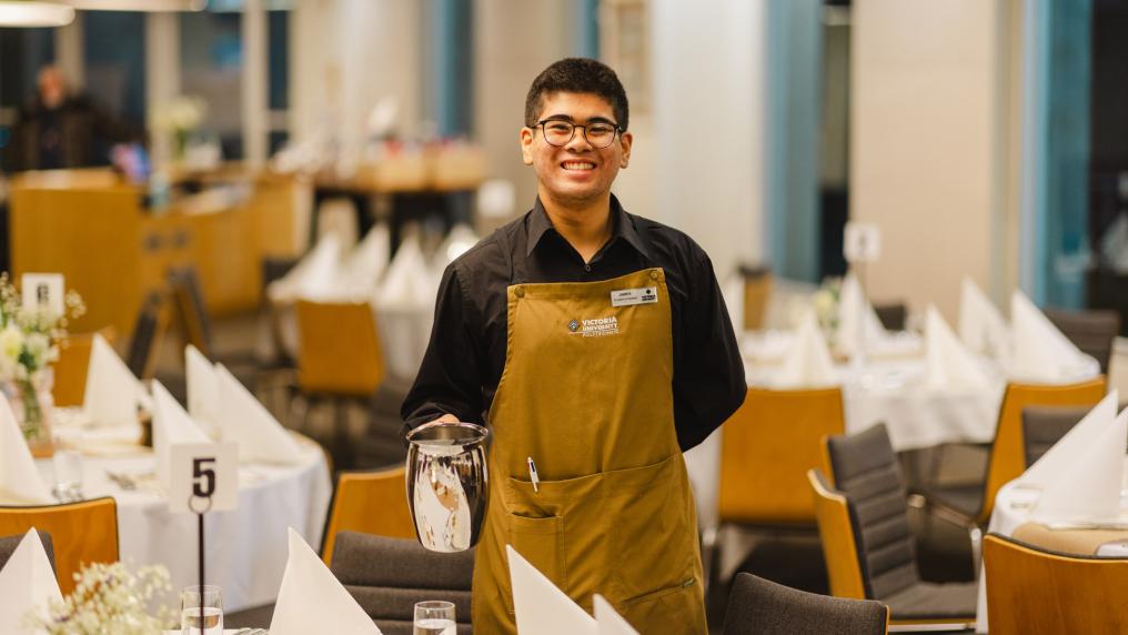 A student in an apron holding a jug of water, smiling. The student is tanding in an empty restaurant, surrounded by decorated tables.