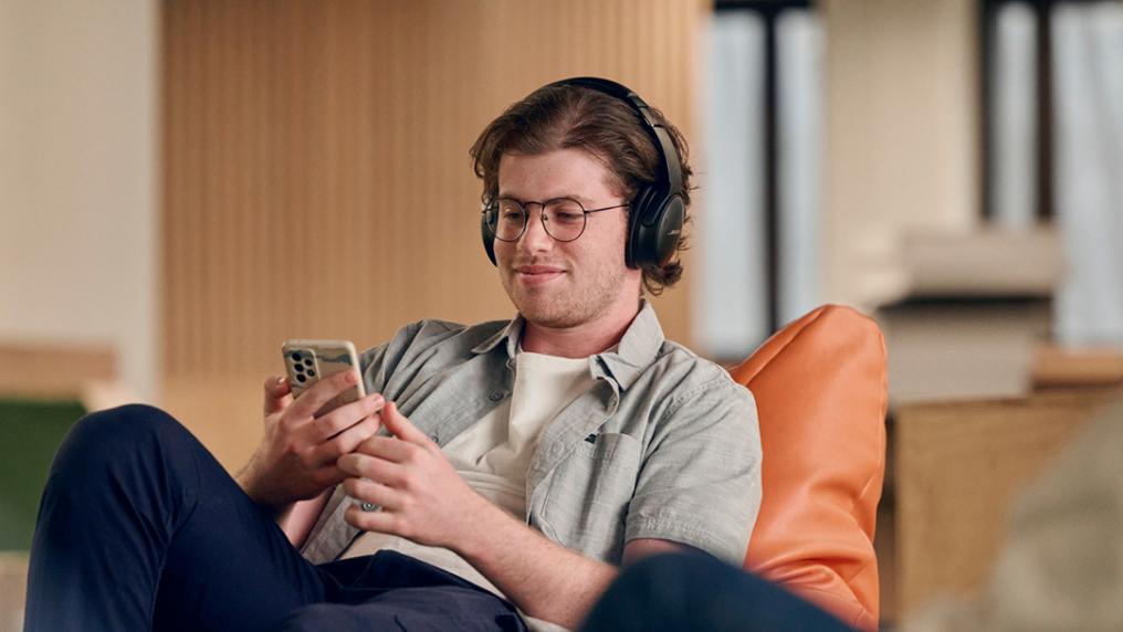 A student sits on a bean bag, with headphones on, holding a mobile phone.