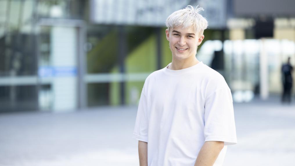 A young person with white-blond hair in a university courtyard smiles happily at the camera