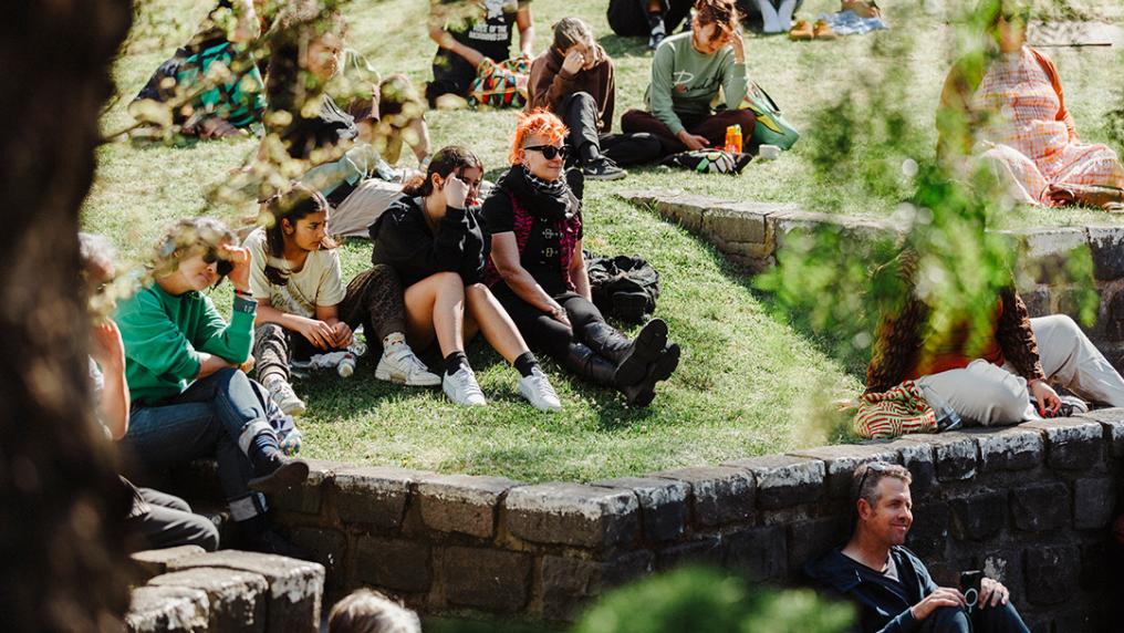 A group of young adults in casual clothes watch a show from a grassy bank