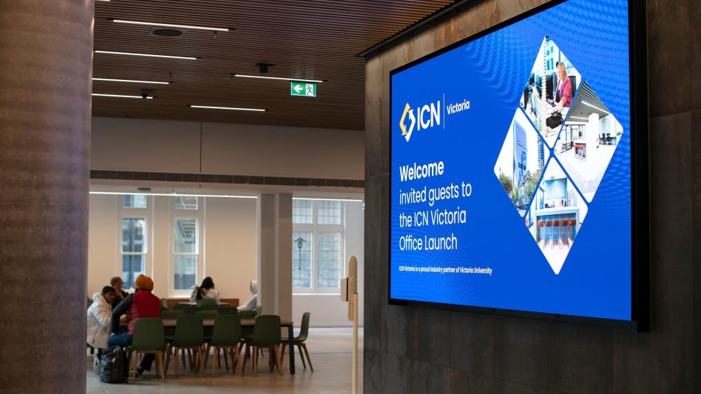 Looking into an office from the hallway. In the hallway is a large screen that says "ICN Victoria. Welcome invited guests to the ICN Victoria Office Launch"