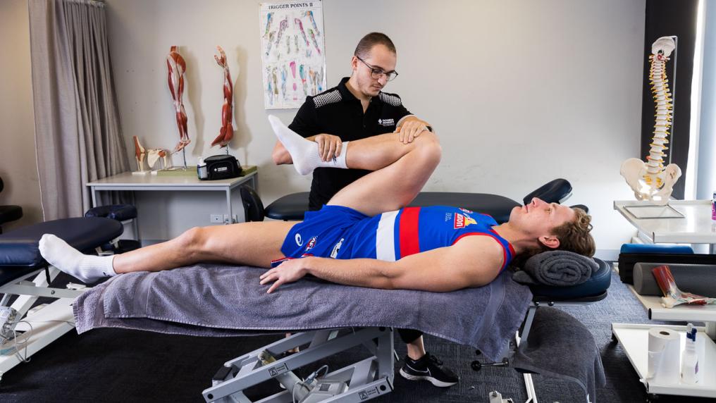 A person in a black & white uniform massages a man wearing a blue and white sport uniform in a room with anatomical posters and models
