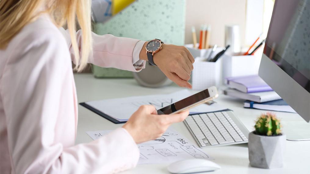 A person at a desktop computer looks at their watch while holding their phone.