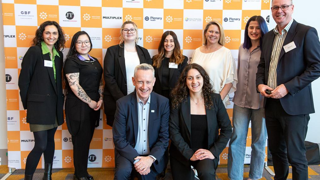 A group of young women and two middle-aged men in suits pose against a wall of yellow and white logos