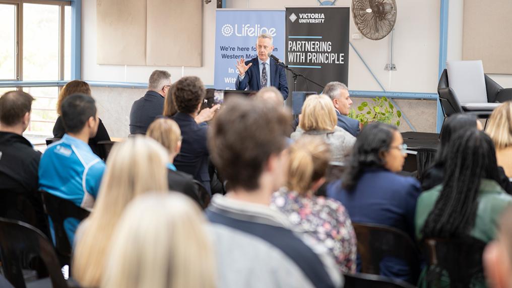 a middle aged man in a suit talks to roomful of seated people, with Lifeline and Victoria University banner behind him that reads 'Partnering with Principle'