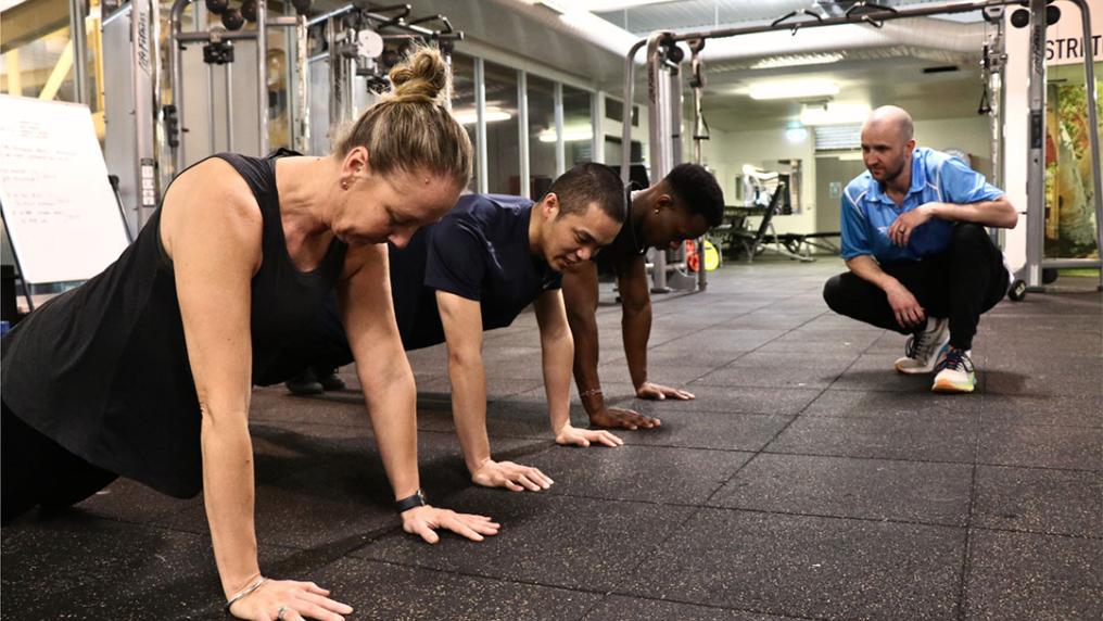 Three people in Footscray Park doing push ups, while a personal trainer encourages them.