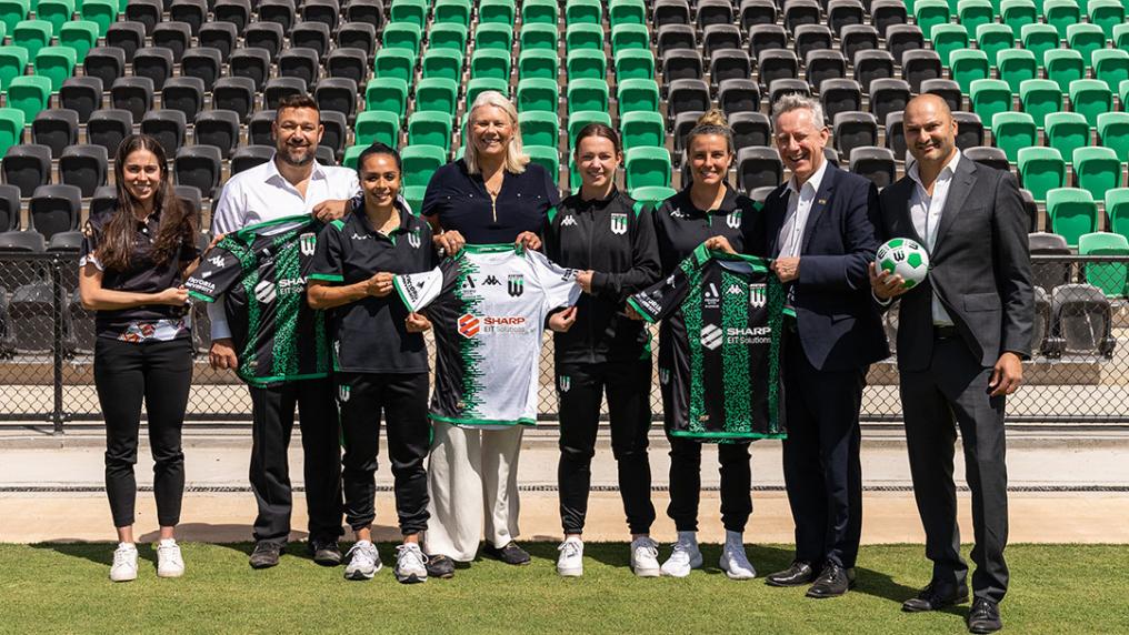 A group of women in sports uniforms and men in professional clothing pose with green sports uniforms and soccer balls in a sport stadium