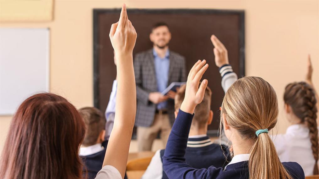 A secondary teacher stands at the front of a classroom, with students in uniforms raising hands to answer questions.
