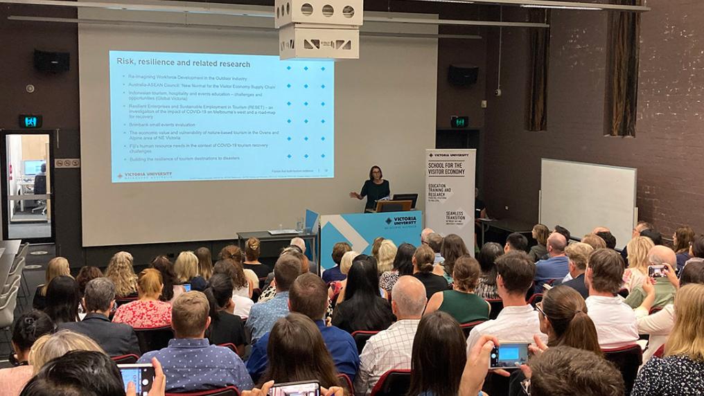 A full conference room with attention towards a presenter in front of a screen