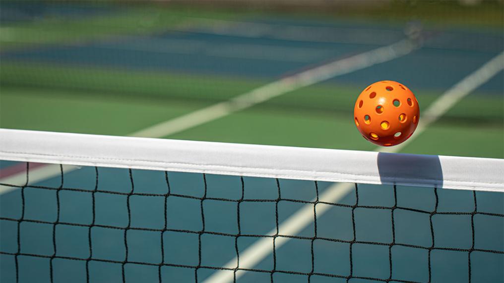 Orange pickleball sailing over net.