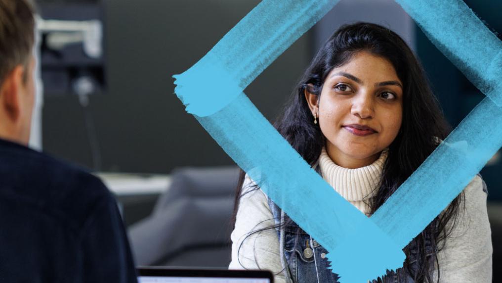 A female student sits in front of a person with a laptop, with the blue branded VU diamond featured around her head.