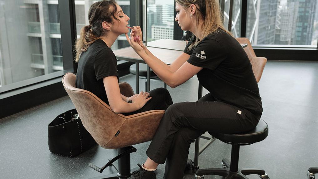 A young woman in a Victoria University uniform applies a customer's make up in a modern, highrise room