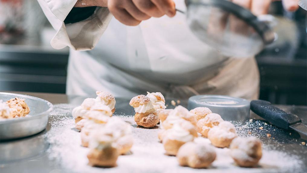 A chef sprinkles sugar through a sieve onto pastry desserts.