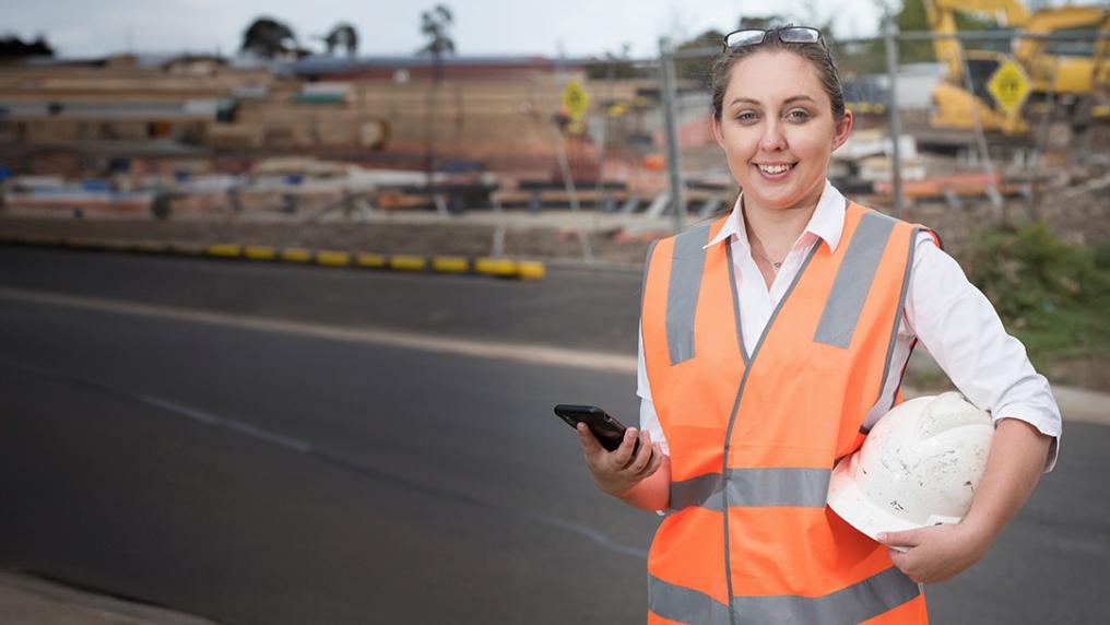 A young woman in a high-vis workers vest holds a hardhat & phone on a construction site