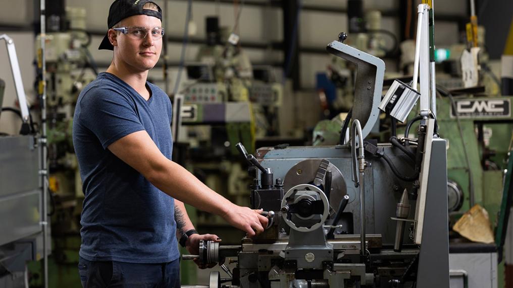 A young man in safety googles operates a large industrial machine in a well-equipped workshop