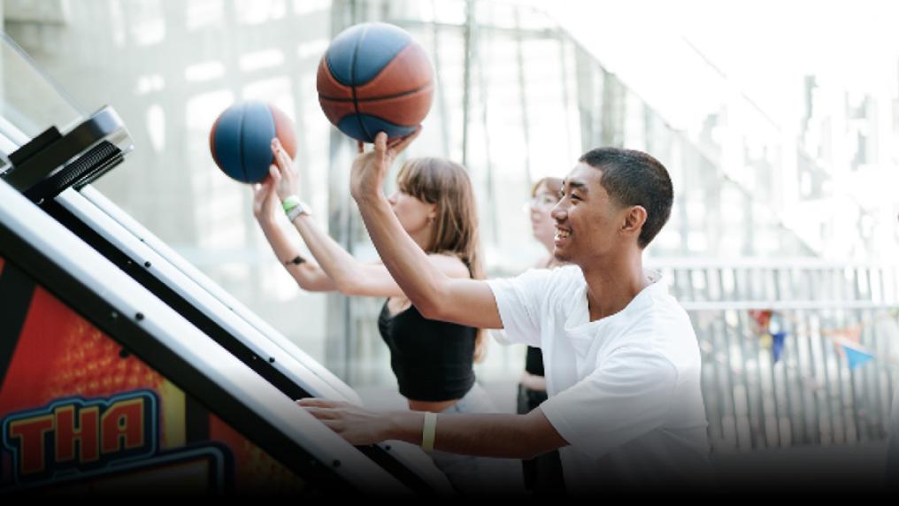 Two students about to throw a basketball into a basketball arcade machine. One student is watching in the background.