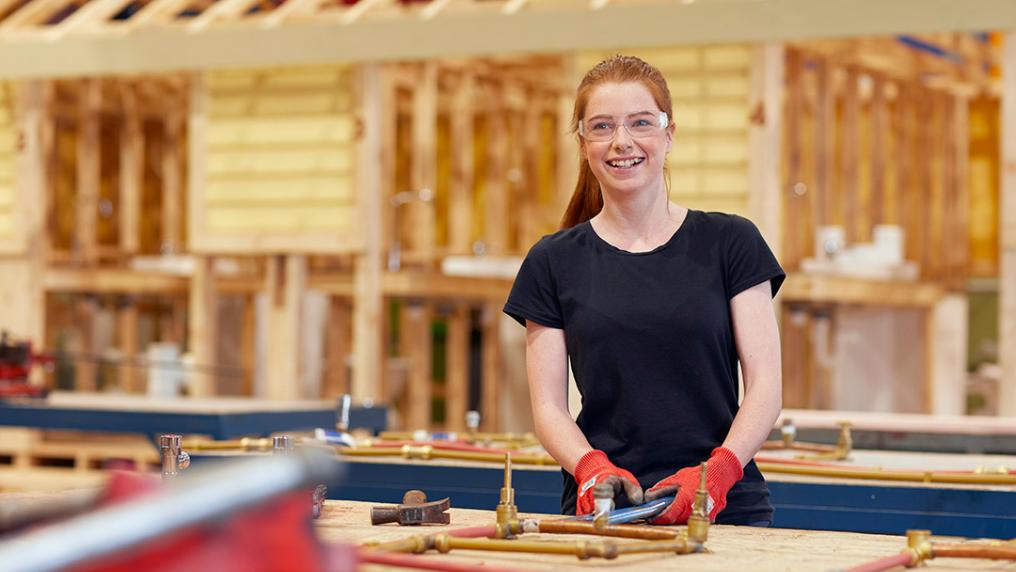 A smiling young woman wears safety goggles and gloves while working in a large carpentry workshop