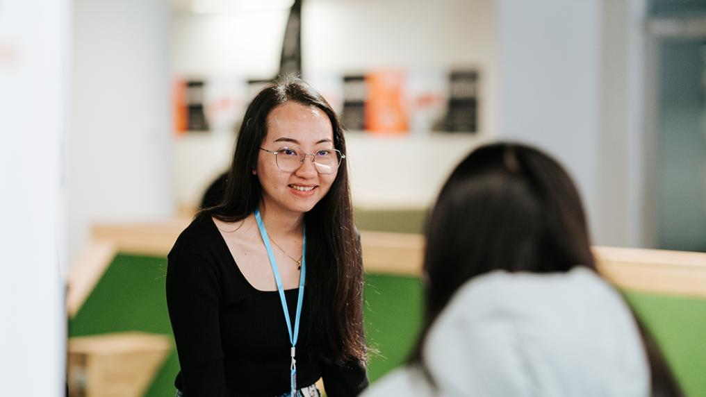 A young woman with a lanyard talks to a student in a bright, modern room