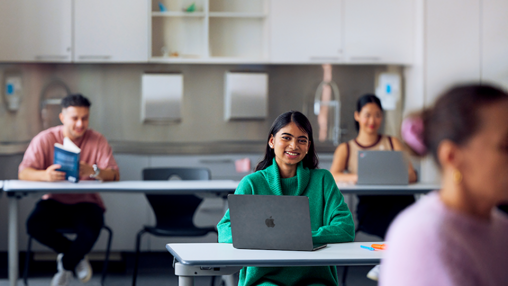 A VU student with a laptop in a classroom, smiling.
