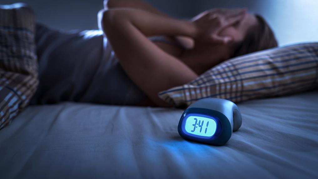 A woman in bed with her hands over her face. A clock reads 3:41.