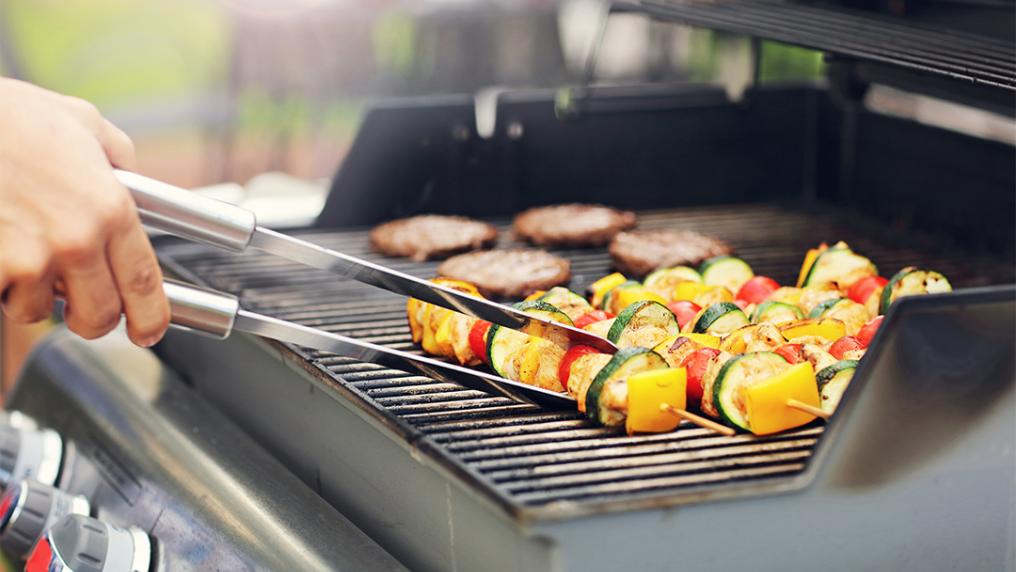 Tongs rearranging veggie skewers over a BBQ.