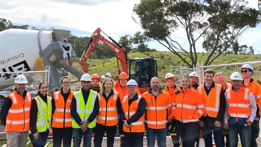 A group of 16 people in hi vis vests smile at the camera, with the trench trial site in background of the photo