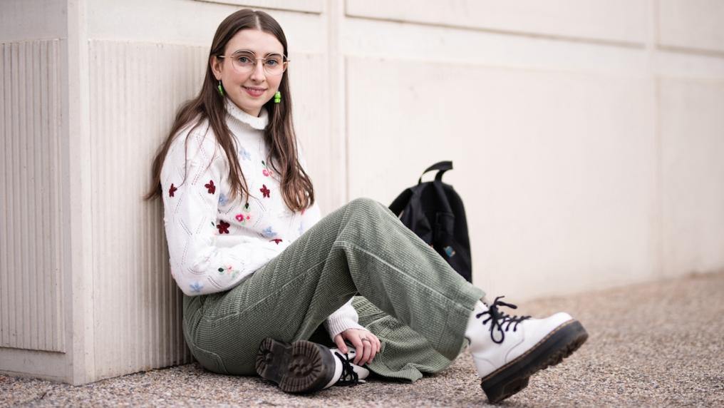 A student wearing glasses and a white sweater with embroidered flowers, sitting casually against a wall with a backpack next to her.