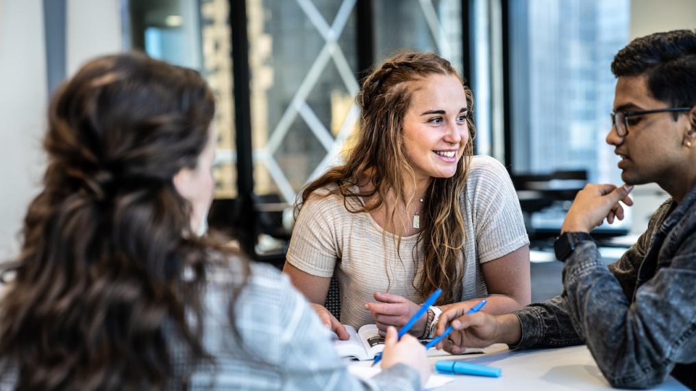 Students engaged in a discussion at a table, with one student smiling brightly as she listens.