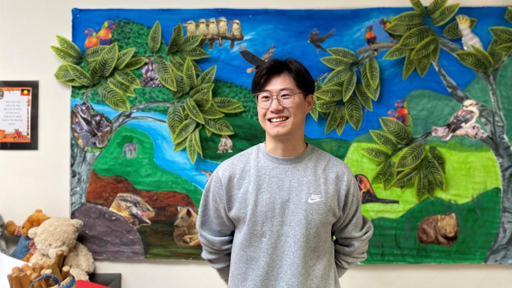 Jarry Shen smiles at the camera. He is standing in front of a fabric art installation at Werribee Childrens Centre