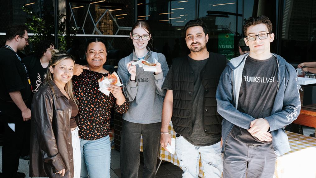 A group of young adults smile in the sun in front of a modern glass window